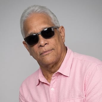 Senior man with white hair wearing dark sunglasses and a pink striped shirt against a gray background