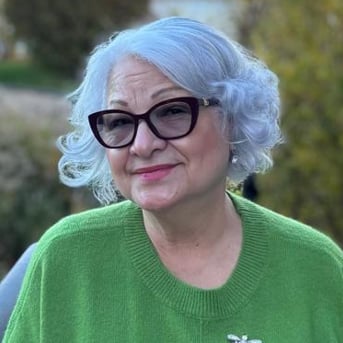 Older woman with short white hair and black-framed glasses wearing a green sweater outdoors