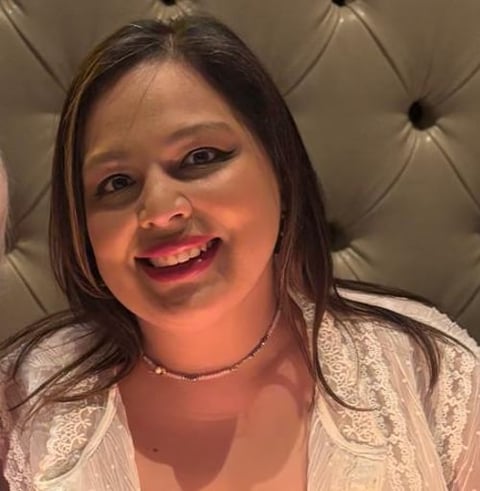Smiling woman with dark hair, wearing a beige lace top and pearl necklace against a tufted beige background